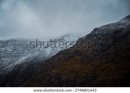 A dramatic mountain landscape showcasing a snow‑capped peak rising sharply against a moody blue sky. In the foreground, a rugged, golden‑brown hillside dotted with sparse vegetation leads to two tall electrical transmission towers with multiple power lines stretching across the scene. The contrast highlights the raw natural beauty of the highland terrain juxtaposed with industrial infrastructure.