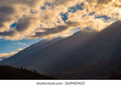 Dramatic mountain landscape with sun rays piercing through clouds, highlighting the rugged terrain under a dynamic sky.