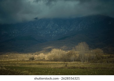Dramatic mountain landscape under stormy clouds with leafless trees and rural fields in late autumn. Moody natural scenery with dark forested hills and contrasting light in the valley, creating a cine
