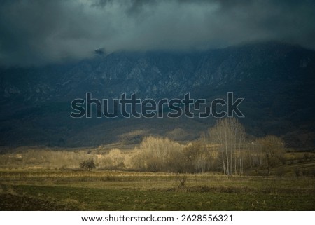 Dramatic mountain landscape under stormy clouds with leafless trees and rural fields in late autumn. Moody natural scenery with dark forested hills and contrasting light in the valley, creating a cine