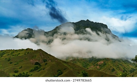 Dramatic mountain peak surrounded by thick mist and rolling green hills under a blue cloudy sky, creating breathtaking tropical highland scenery and natural landscape.