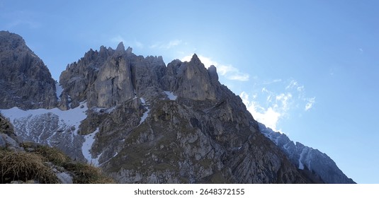 A dramatic mountain range with jagged peaks silhouetted against a bright, clear blue sky, hinting at the rising or setting sun behind. Patches of snow visible on the slopes.