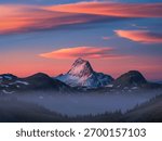 A dramatic mountain range under a vibrant sunset sky with wispy clouds, featuring a prominent snow-capped peak above a misty valley.