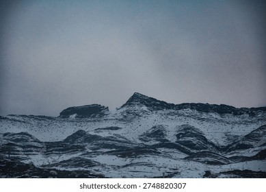 Dramatic mountain scenery of snow‑capped peaks framing a winding road through rugged highland terrain under an atmospheric sky with misty fog. The alpine landscape showcases a natural backdrop of wilderness photography with elevation view and outdoor adventure setting.