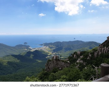 Dramatic mountain summit view overlooking a vast coastal landscape with green forested mountains, rocky outcrops in the foreground, and the blue sea stretching to the horizon with ships and islands. A coastal town is nestled in the valley below.