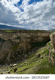 Dramatic mountain valley landscape with deep rocky gorge, flowing river, green meadows, snow-capped peaks, and dramatic cloudy sky.