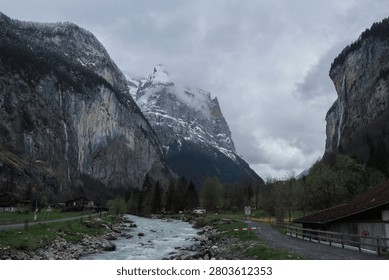 A dramatic mountain valley landscape featuring a flowing river, dense forest, and snow-covered peaks under a cloudy sky