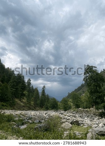 A dramatic mountain valley scene under a brooding sky. Dark, heavy storm clouds swirl overhead, creating a moody atmosphere and soft diffused light. Below, a rocky riverbed stretches through the center of the landscape, surrounded by lush green trees and pine-covered slopes on both sides. In the distance, misty hills fade into the horizon, adding depth and a sense of wilderness. The contrast between the rugged rocks, dense forest, and turbulent sky gives the image a calm yet powerful natural beauty.