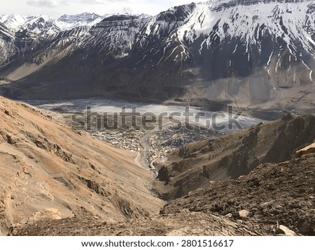 Dramatic mountain valley with snow covered peaks and rugged terrain, breathtaking Himalayan landscape overlooking a remote settlement.