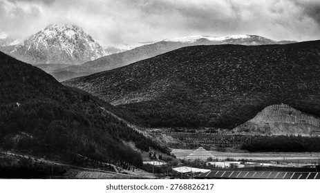A dramatic mountain view under heavy clouds shows layered hills and distant snowy peaks. In black and white, the prayer flags blend into texture, while contrast and light emphasize the vast scale and quiet mood of the land.
