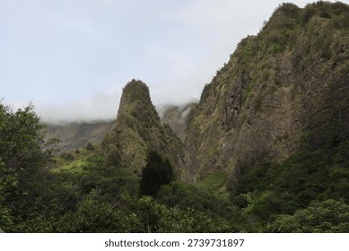 A dramatic, narrow mountain peak rises sharply above dense tropical greenery, surrounded by mist and rugged cliffs. Lush vegetation and low clouds create a mysterious, atmospheric landscape.