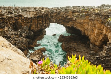 A dramatic natural arch carved into the rocky cliffs, with turquoise waves crashing beneath, surrounded by rugged terrain and vibrant coastal plants - Powered by Shutterstock