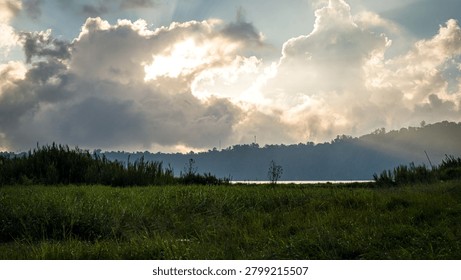 A dramatic natural landscape with rays of sunlight breaking through thick clouds over a vast green meadow on the edge of a lake.