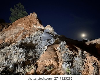 Dramatic night landscape featuring illuminated rocky formations under a dark blue sky with a bright full moon. The rugged sandstone ridge and textured terrain are highlighted by artificial lighting, creating strong contrasts between warm earth tones and cool night shadows. Wild vegetation grows among the rocks, adding depth and detail to the natural composition. The scene conveys a sense of wilderness, mystery, and remote outdoor exploration, ideal for nature, adventure, geology, and night photography projects.