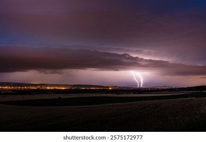 A dramatic night landscape featuring a vivid lightning strike over a distant illuminated city, under a moody cloudy sky.