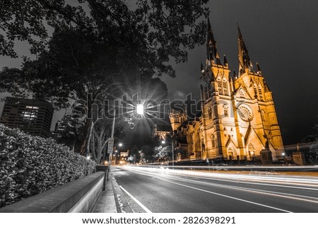 Dramatic night view of an illuminated Gothic cathedral beside a city street, featuring bright long exposure light trails from passing traffic. The composition uses a wide street-level perspective with dark tree silhouettes, urban buildings, road lines, and glowing architectural details creating strong contrast. The selective color style highlights warm golden light on the historic church against a mostly black and white cityscape, giving the image a cinematic, atmospheric, and timeless mood. Ideal for travel, tourism, architecture, religion, urban night photography, city promotion, editorial design, cultural heritage content, posters, brochures, websites, and commercial backgrounds.