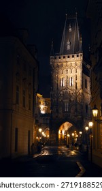 Dramatic night view of the Powder Tower (Prašná brána) in Prague, Czech Republic, illuminated by warm glowing street lights, moody and atmospheric evening. 