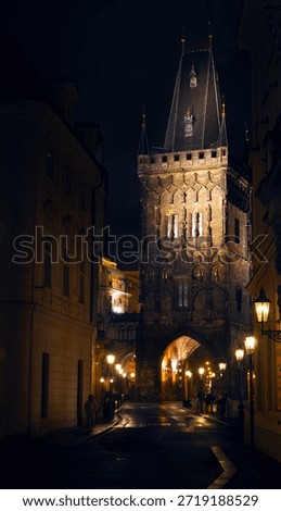 Dramatic night view of the Powder Tower (Prašná brána) in Prague, Czech Republic, illuminated by warm glowing street lights, moody and atmospheric evening. 