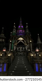 A dramatic nighttime scene featuring a grand, castle-like structure with gothic architectural elements. The building is illuminated with vibrant purple, green, and warm golden lights, creating a mystical and fantasy-like atmosphere. A central staircase leads up to an ornate entrance framed by pointed arches, decorative lanterns, and intricate details. The dark sky enhances the glowing lights, giving the scene a magical, almost fairytale ambiance.