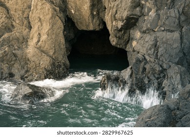 A dramatic ocean cave opens toward vibrant turquoise water, with seawater flowing and cascading gently from the rugged rock formations along the cave walls. The textured stone surfaces contrast beautifully with the clear blue-green water, creating a peaceful and immersive coastal landscape. Natural light filters into the cavern, highlighting the movement of the water and intricate details of the surrounding rocks while adding depth and atmosphere to the scene.