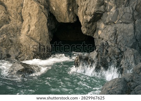 A dramatic ocean cave opens toward vibrant turquoise water, with seawater flowing and cascading gently from the rugged rock formations along the cave walls. The textured stone surfaces contrast beautifully with the clear blue-green water, creating a peaceful and immersive coastal landscape. Natural light filters into the cavern, highlighting the movement of the water and intricate details of the surrounding rocks while adding depth and atmosphere to the scene.