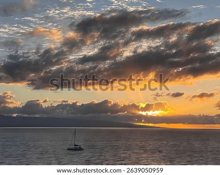 Dramatic ocean sunset off the coast of Maui, Hawaii, with a sailboat floating peacefully on calm waters beneath vibrant clouds illuminated by golden and orange evening light.