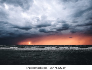 Dramatic ocean view with stormy clouds and a vibrant orange sunset on the horizon. Capturing the contrast between dark, moody skies and the warm glow of the setting sun over the sea.