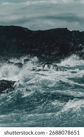 Dramatic ocean waves crash against rugged rocky cliffs under a cloudy sky.