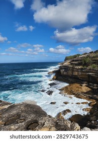 Dramatic ocean waves crash against rugged sandstone cliffs along the scenic Bondi to Coogee coastal walk in Sydney, Australia. Clear blue waters, sea foam, and bright skies create a beautiful natural landscape, perfect for coastal adventure travel.