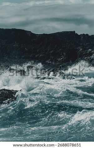 Dramatic ocean waves crash against rugged rocky cliffs under a cloudy sky.
