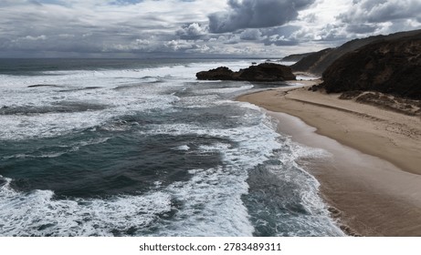 Dramatic ocean waves hit a sandy shore and rocky cliffs under a cloudy sky.