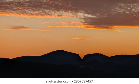Dramatic orange and pink sunset sky over a dark silhouetted mountain ridge, with scattered clouds catching the last golden light of evening.