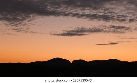 Dramatic orange and pink sunset sky over a dark silhouetted mountain ridge, with scattered clouds catching the last golden light of evening.