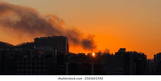 A dramatic orange sunset illuminates the city skyline, with dark buildings silhouetted against the fiery sky as smoke or clouds drift across the horizon. - Powered by Shutterstock