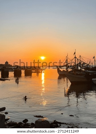 A dramatic orange sunset reflects on the calm harbor water, highlighting traditional boats moored along the pier. Peaceful coastal life scene