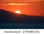 Dramatic orange sunset with the sun setting behind a dark mountain ridge and a calm sea foreground — panoramic minimalist landscape for backgrounds and travel content.