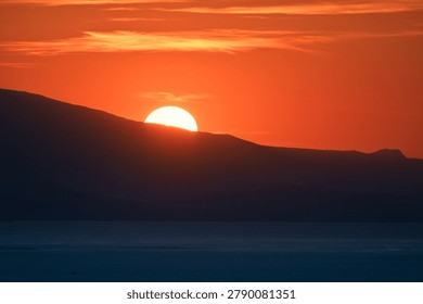 Dramatic orange sunset with the sun setting behind a dark mountain ridge and a calm sea foreground — panoramic minimalist landscape for backgrounds and travel content.