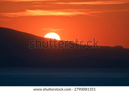 Dramatic orange sunset with the sun setting behind a dark mountain ridge and a calm sea foreground — panoramic minimalist landscape for backgrounds and travel content.