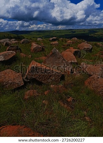 A dramatic outdoor landscape featuring large , angular reddish-brown granite boulders scatterd across lush grreen grassy terrain under a partly cloudy sky with dramatic cumulus clouds in the background