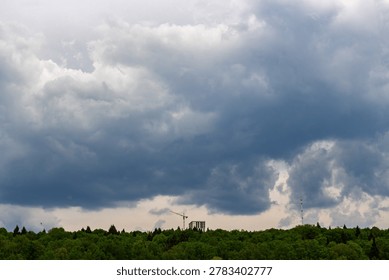 Dramatic overcast spring sky filled with heavy storm clouds hanging above a lush green forest canopy featuring a distant construction site with a crane and a tall communication tower