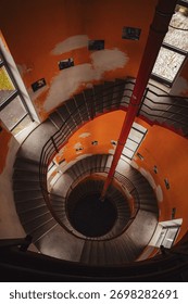 Dramatic overhead view looking down a vintage spiral staircase in a tower, with peeling orange walls and strong geometric lines, emphasizing infinite depth and architectural detail.
