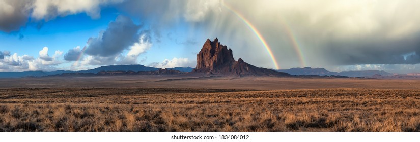 Dramatic panoramic landscape view of a dry desert with a mountain peak in the background. Dramatic Sunrise with Rainbow Artistic Render. Taken at Shiprock, New Mexico, United States.