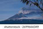 Dramatic panoramic view of an active volcano emitting smoke and ash, with low clouds and mist enveloping its base against a deep blue sky.