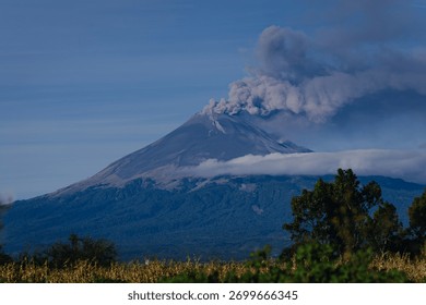 Dramatic panoramic view of an active volcano emitting smoke and ash, with low clouds and mist enveloping its base against a deep blue sky.