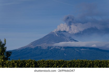 Dramatic panoramic view of an active volcano emitting smoke and ash, with low clouds and mist enveloping its base against a deep blue sky.