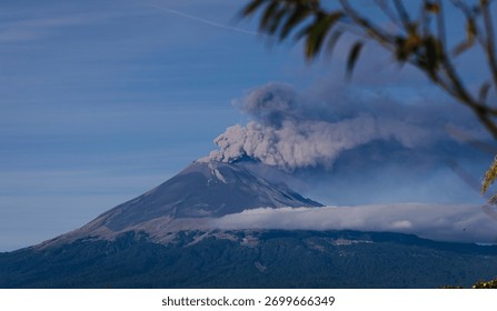 Dramatic panoramic view of an active volcano emitting smoke and ash, with low clouds and mist enveloping its base against a deep blue sky.