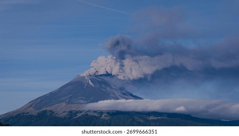 Dramatic panoramic view of an active volcano emitting smoke and ash, with low clouds and mist enveloping its base against a deep blue sky.