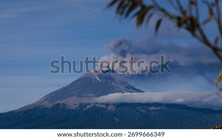 Dramatic panoramic view of an active volcano emitting smoke and ash, with low clouds and mist enveloping its base against a deep blue sky.