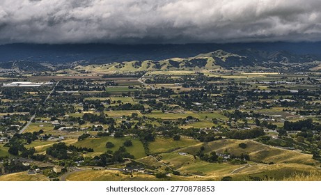 A dramatic panoramic view from a grassy hillside overlooking a vast green valley with patchwork fields, a sprawling town, and distant mountains under a moody, cloudy sky.