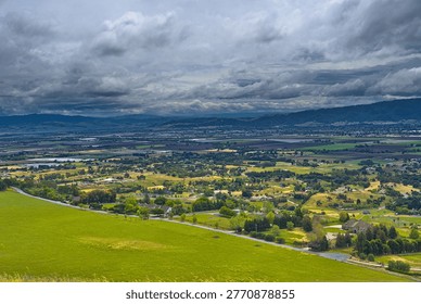 A dramatic panoramic view from a grassy hillside overlooking a vast green valley with patchwork fields, a sprawling town, and distant mountains under a moody, cloudy sky.